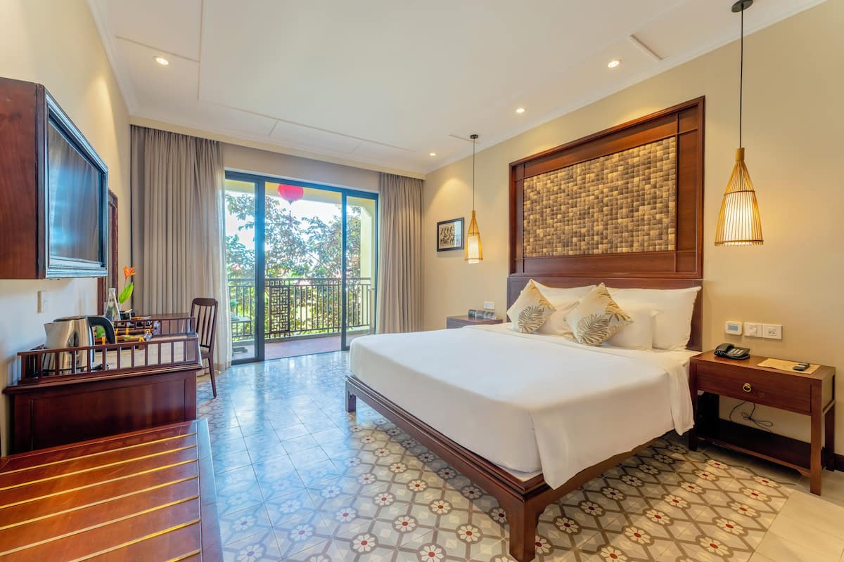 Ein Hotelzimmer mit einem großen Bett, einem Schreibtisch, einem Sessel, einem Fernseher, einem Balkon mit Vorhängen und einem gemusterten Boden.