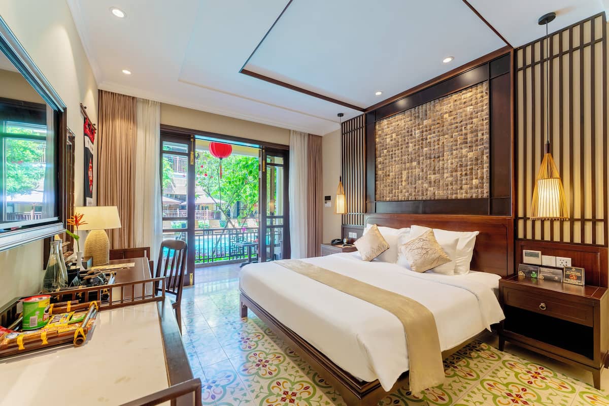 Ein Hotelzimmer mit einem großen Bett, einem Schreibtisch mit Stuhl, einem Fernseher und einem Balkon mit Blick auf Grünflächen.