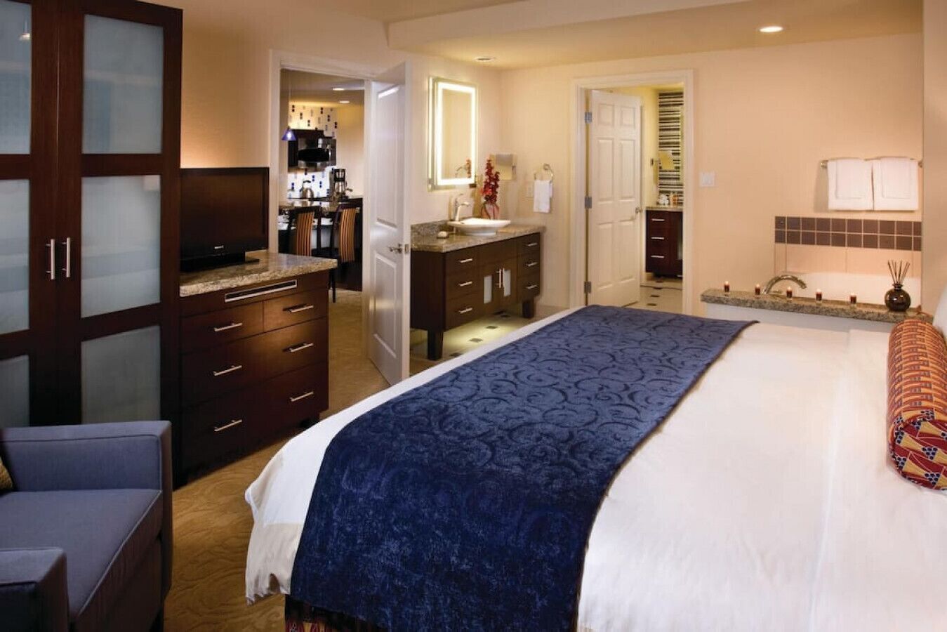 A hotel room with a large bed, a desk, a chair, a wardrobe, a bathroom with a mirror, and a kitchenette.
