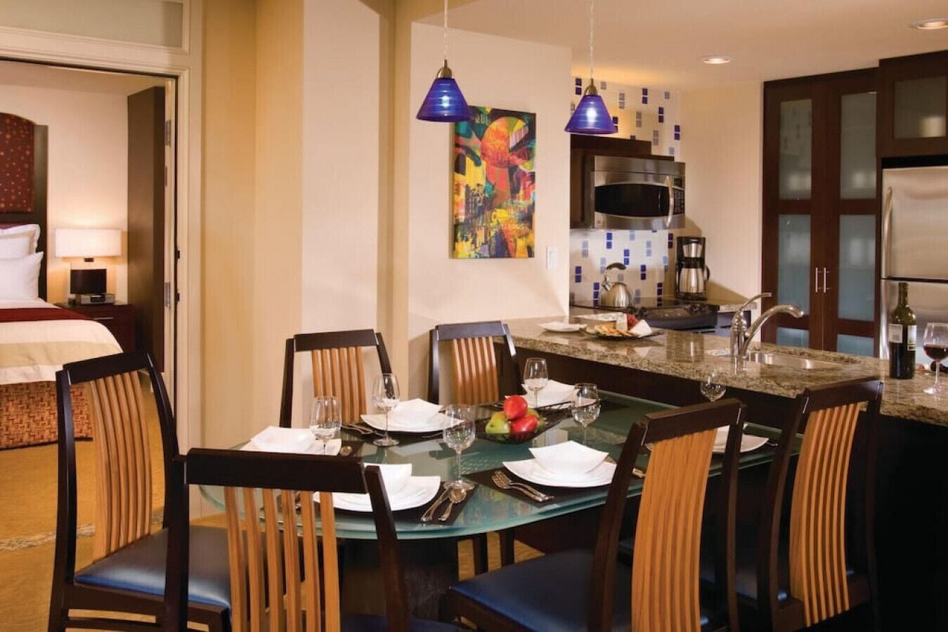 Un comedor de hotel con una mesa de cristal preparada para seis personas, rodeada de sillas de madera. Se ve una cocina con microondas y estufa al fondo.