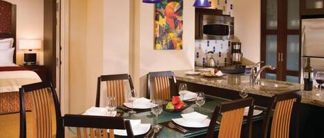 Un comedor de hotel con una mesa de cristal preparada para seis personas, rodeada de sillas de madera. Se ve una cocina con microondas y estufa al fondo.