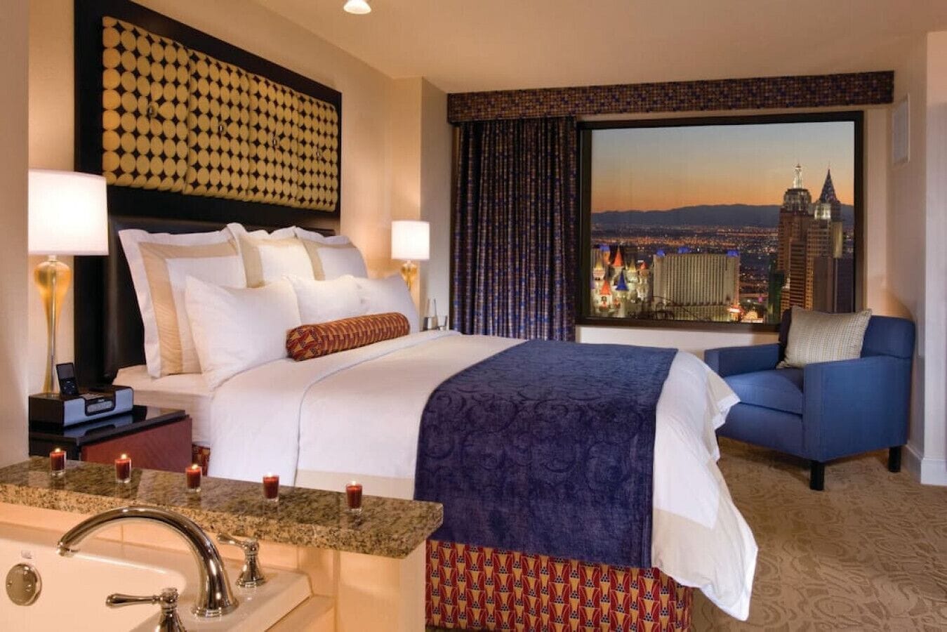 A hotel room with a bed, a desk with a lamp, a chair, a large window with a city view, and a bathroom with a sink and mirror.