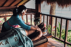 Um deck de madeira com telhado de palha, uma mesa de massagem e vista para o oceano.