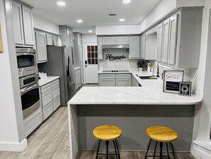 Une cuisine moderne avec des meubles blancs, des appareils en acier inoxydable, un comptoir en marbre et deux tabourets en bois.
