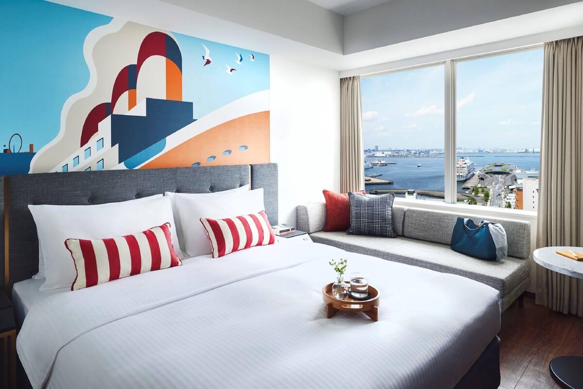 A modern hotel room with a large bed, a view of the city, and a cityscape mural on the wall.
