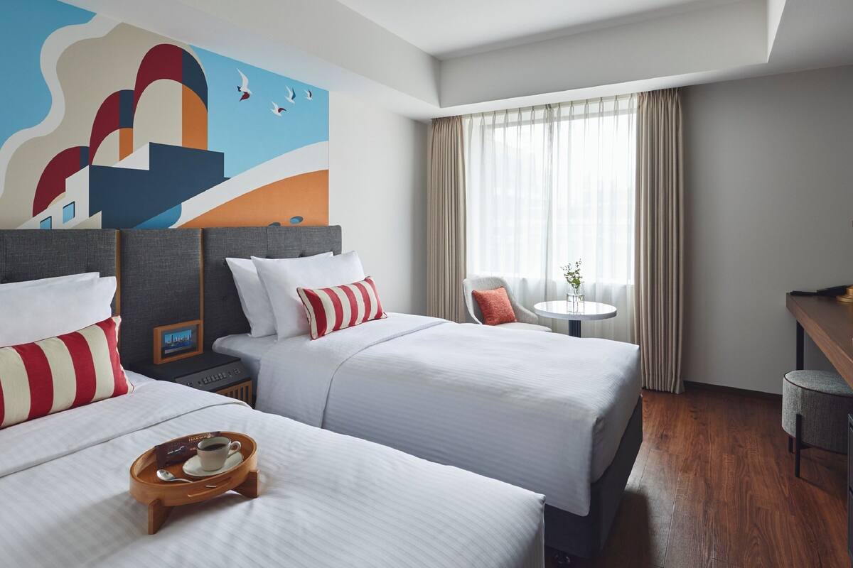 A hotel room with two beds, a small table with a cup and saucer, a chair, and a painting on the wall.
