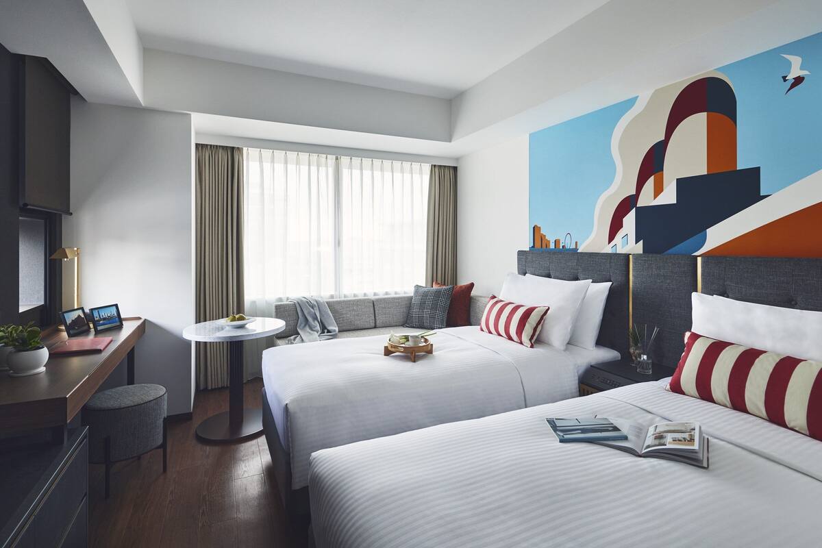 A hotel room with two beds, a desk, a small table, and a large abstract painting on the wall.