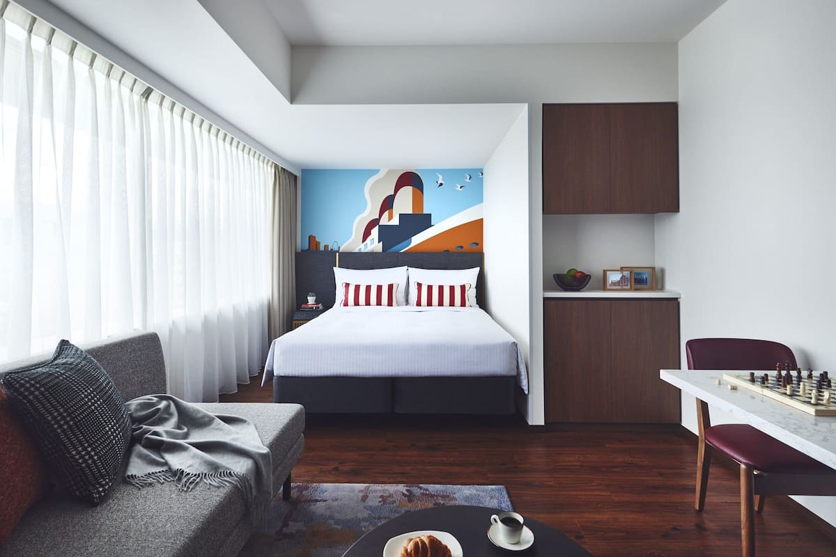 A modern hotel room with a large bed, a sofa, a dining table with chess, and a mural on the wall.