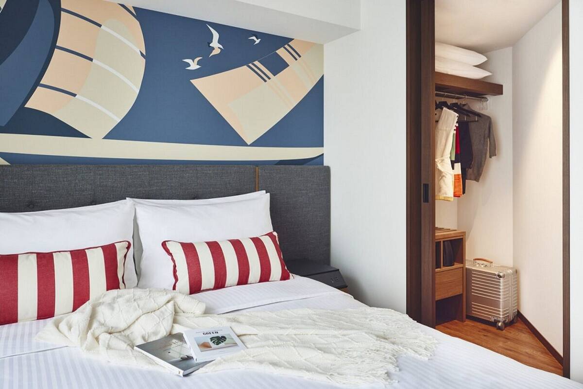 A hotel room with a bed, a wall with abstract art, a wardrobe, and a suitcase.