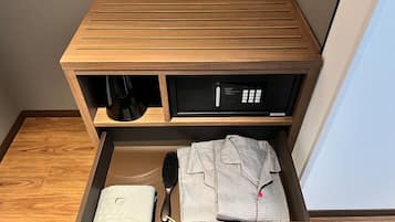 A wooden bedside table with a black electronic device and a drawer containing a white pillow, a black brush, and a patterned shirt.