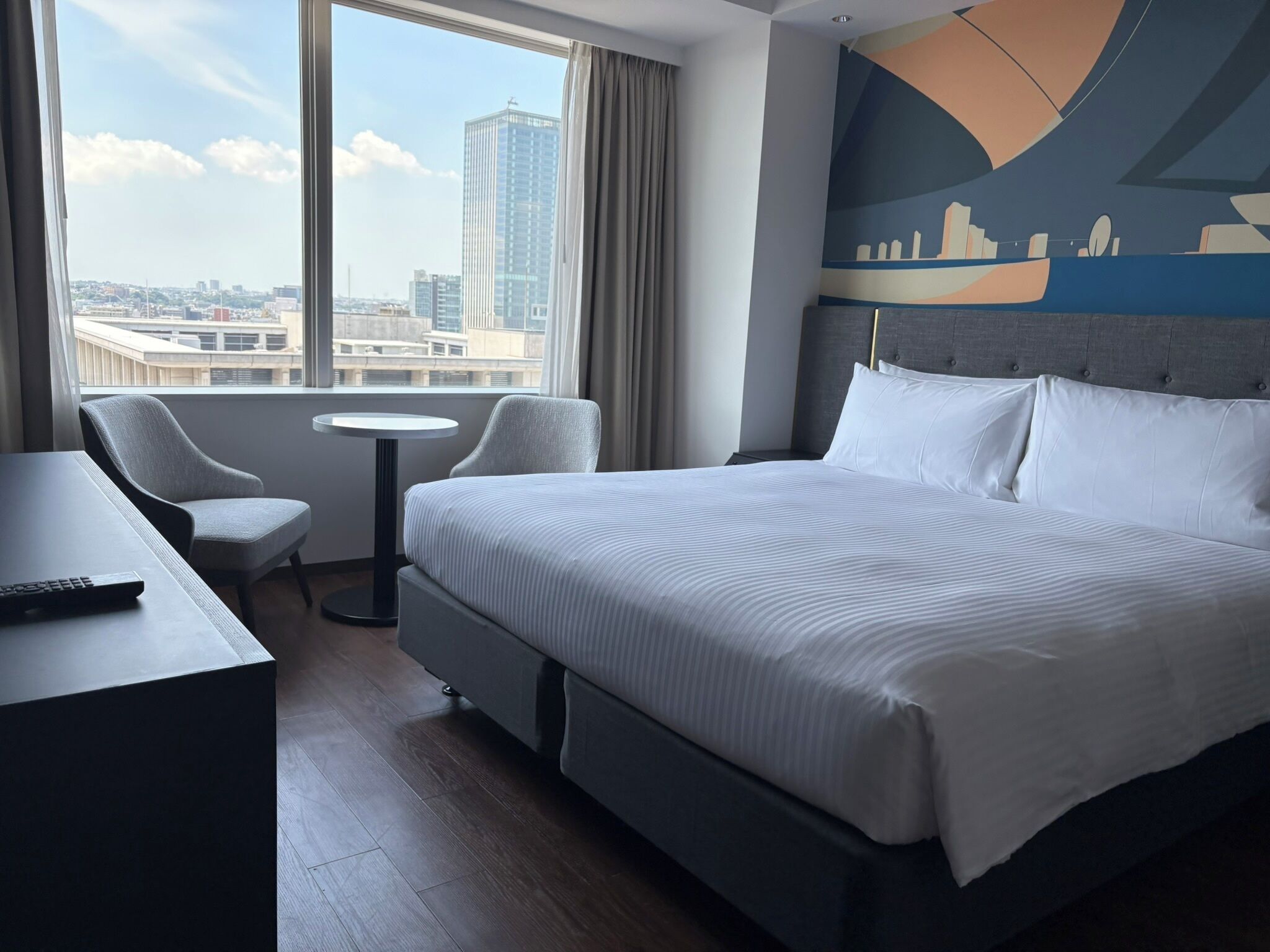 A hotel room with a large bed, a desk, a chair, and a view of the cityscape through the window.