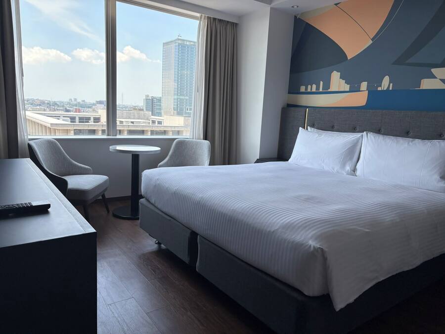 Une chambre d’hôtel avec un grand lit, un bureau, une chaise et une vue sur le paysage urbain par la fenêtre.