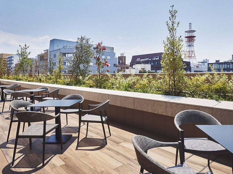 An outdoor seating area with tables and chairs, surrounded by greenery and overlooking a cityscape with modern buildings and a communication tower.