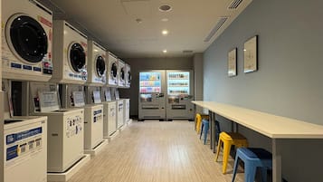 A laundromat with multiple washing machines and dryers, a vending machine, and a long table with stools.