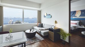 Ein modernes Hotelzimmer mit großem Fenster und Blick auf ein Kreuzfahrtschiff und die Stadtlandschaft. Das Zimmer verfügt über eine Couch, einen runden Couchtisch, einen Beistelltisch mit Lampe sowie einen kleinen Essbereich mit Tisch und Stühlen.
