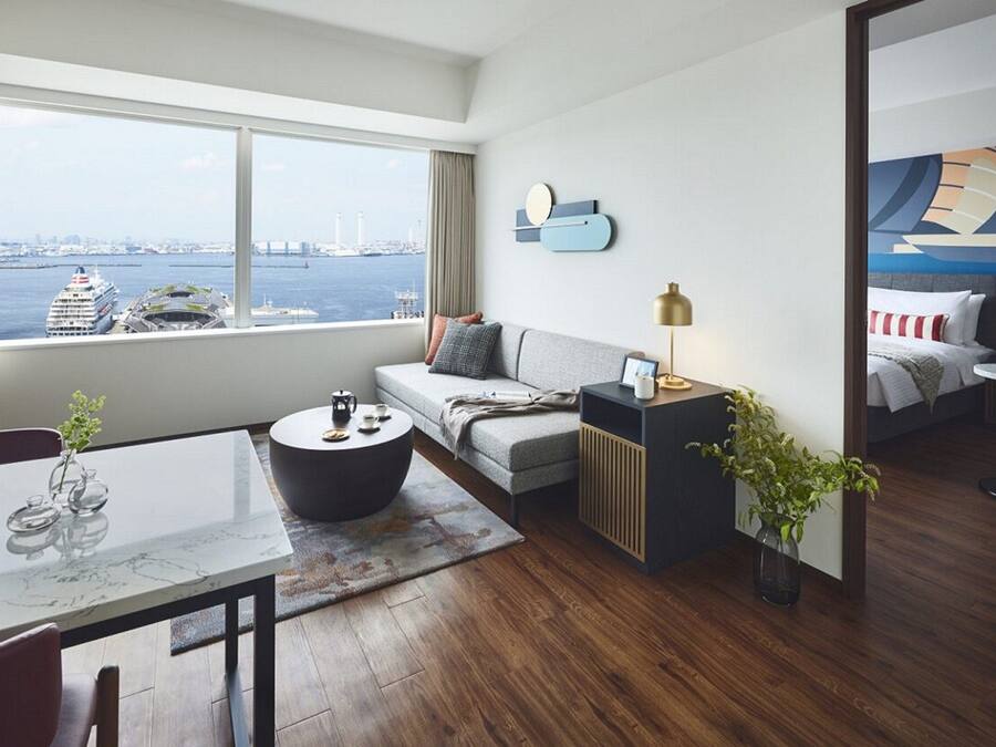 Ein modernes Hotelzimmer mit großem Fenster und Blick auf ein Kreuzfahrtschiff und die Stadtlandschaft. Das Zimmer verfügt über eine Couch, einen runden Couchtisch, einen Beistelltisch mit Lampe sowie einen kleinen Essbereich mit Tisch und Stühlen.