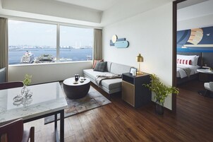 Habitación de hotel moderna con un ventanal que ofrece vistas a un crucero y al horizonte de la ciudad. La habitación cuenta con un sofá, una mesa redonda, una mesita de noche con lámpara y un pequeño comedor con mesa y sillas.