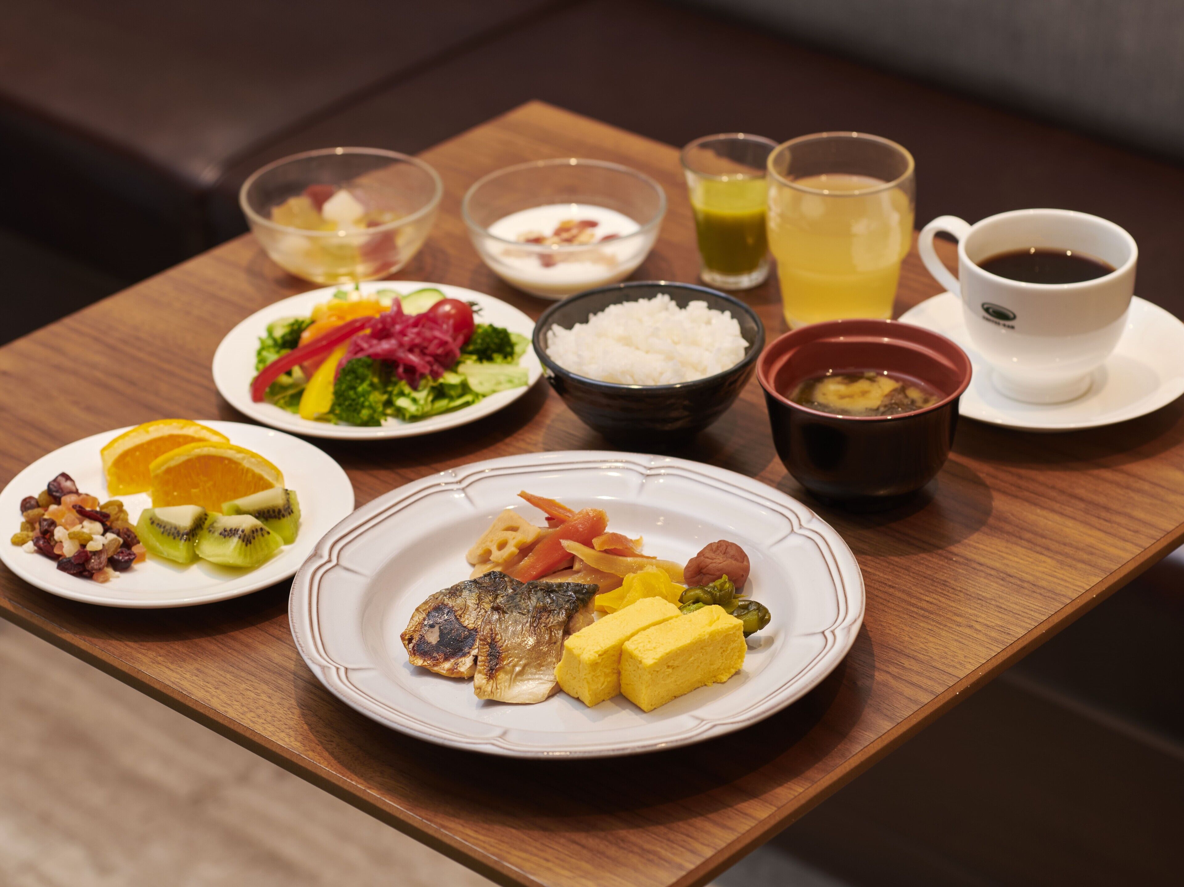 A wooden table with various dishes including fish, vegetables, and rice. There are also bowls of soup and a cup of coffee.