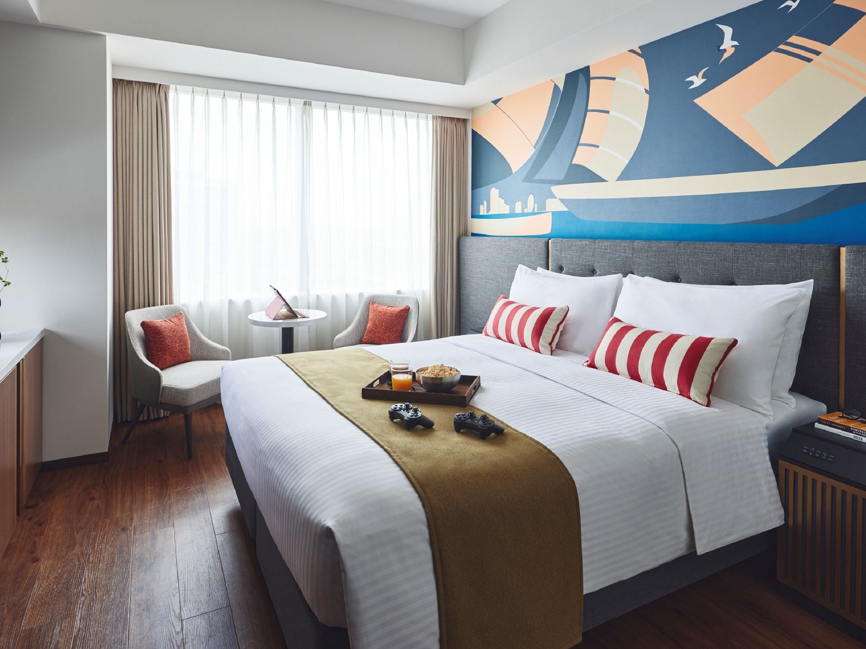 A hotel room with a large bed, a desk, a chair, a window with curtains, and a wall mural.