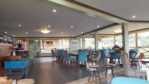 A spacious indoor area with multiple tables and chairs, wooden flooring, and a view of greenery outside.