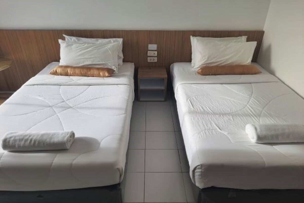 A hotel room with two single beds, each with white bedding and a brown pillow. There is a wooden headboard, a small wooden nightstand with a lamp, and a wall-mounted light switch.