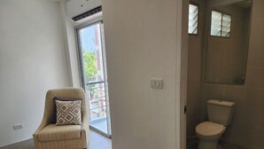 A room with a beige armchair, a pink rug, a white toilet, and a window with a view of greenery outside. - Patong Bay Inn (Patong)