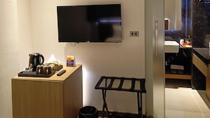 Une chambre d’hôtel moderne équipée d’un téléviseur à écran plat fixé au mur, d’une table de chevet en bois avec une bouilloire et une machine à café, d’une petite poubelle et d’un présentoir en métal noir.