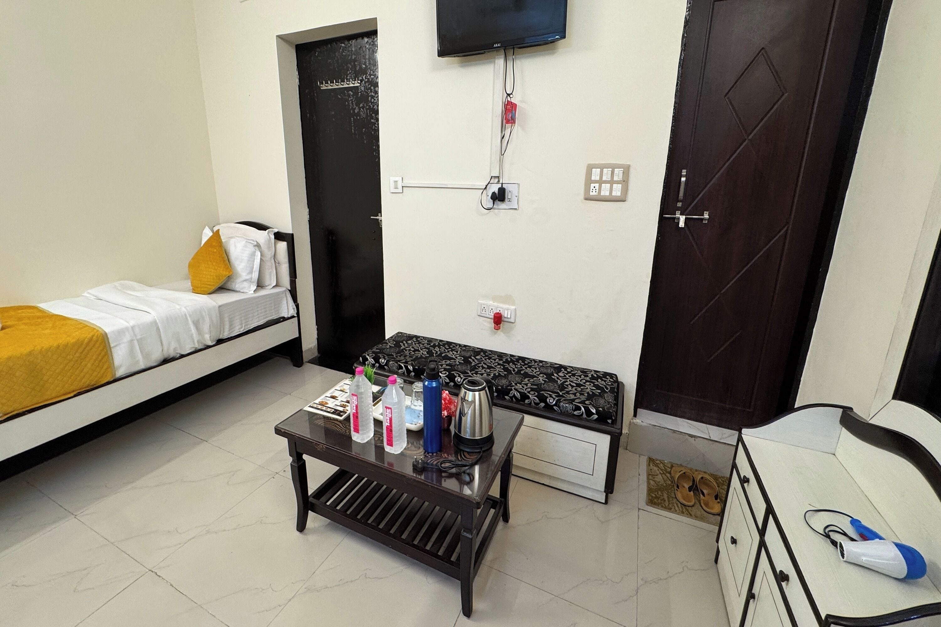 Una habitación pequeña y limpia con una cama, una mesa con botellas de agua, una tetera y un televisor montado en la pared.