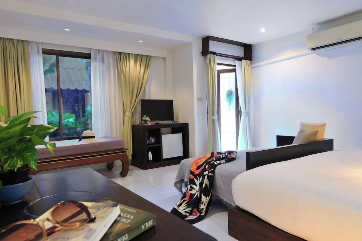 A hotel room with a bed, a TV, a small table with a plant and a book, and a window with curtains.