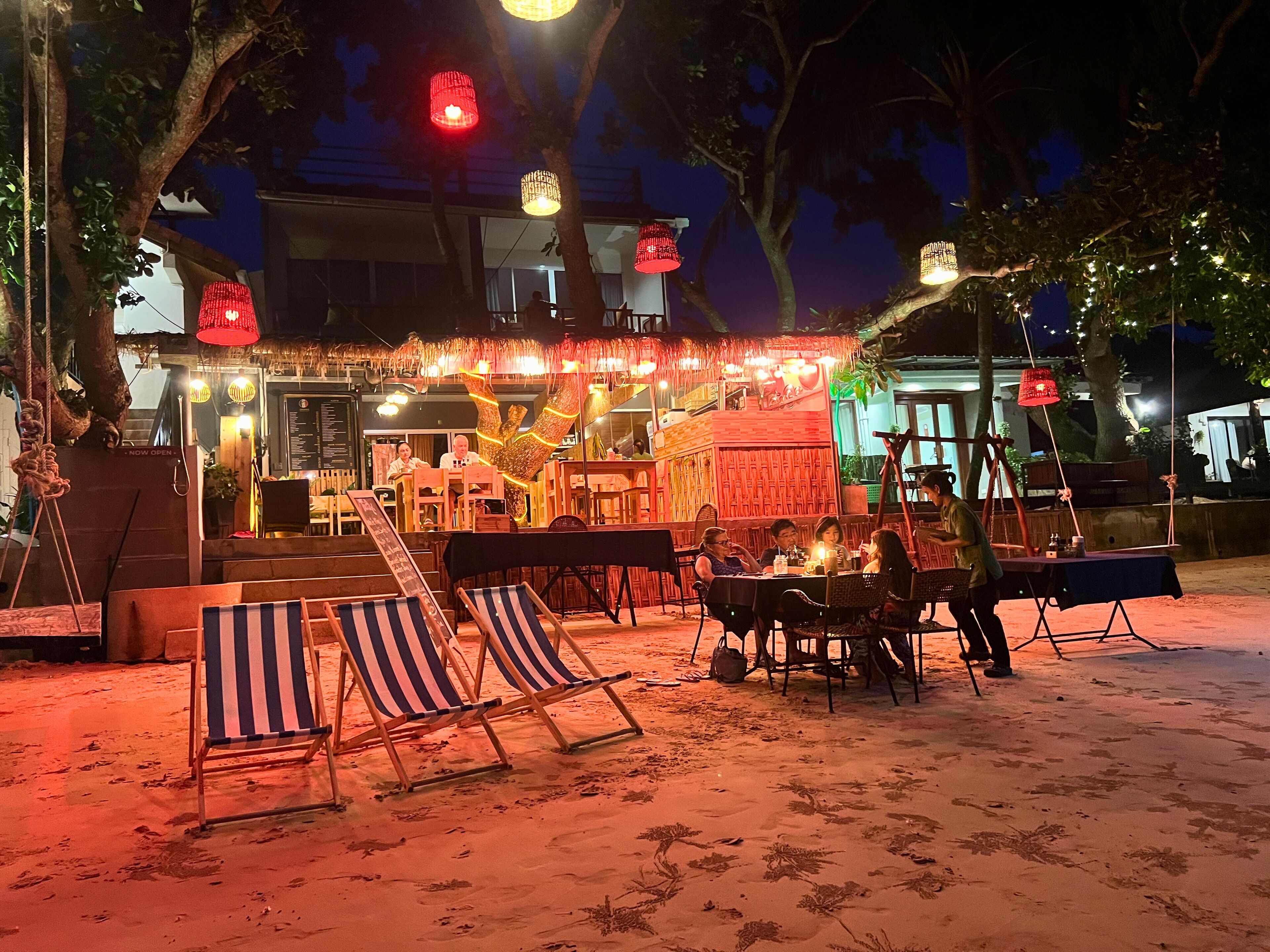 An outdoor dining area with beach chairs and tables, a thatched roof structure, and string lights.