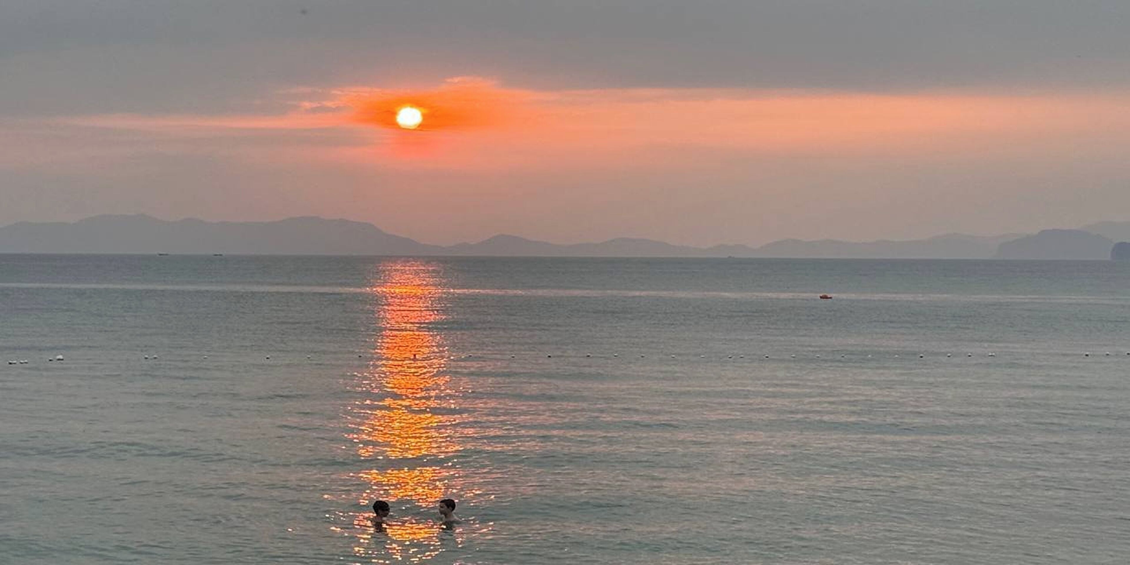 Een zonsondergang boven een rustige zee, met twee mensen die op de voorgrond zwemmen.