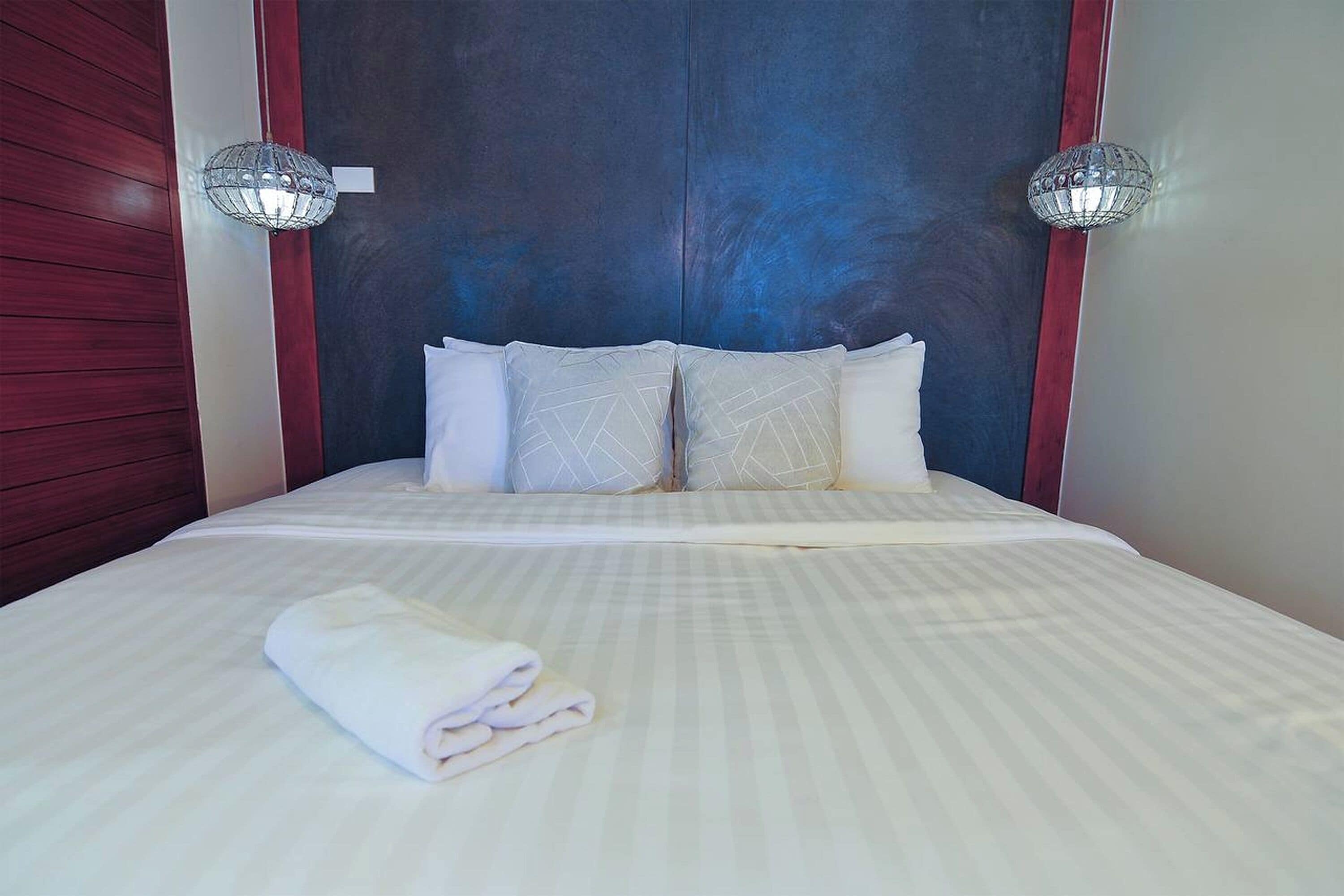 Una cama bien hecha con una colcha a rayas blancas y grises, dos apliques decorativos en la pared y una toalla blanca doblada sobre la cama.