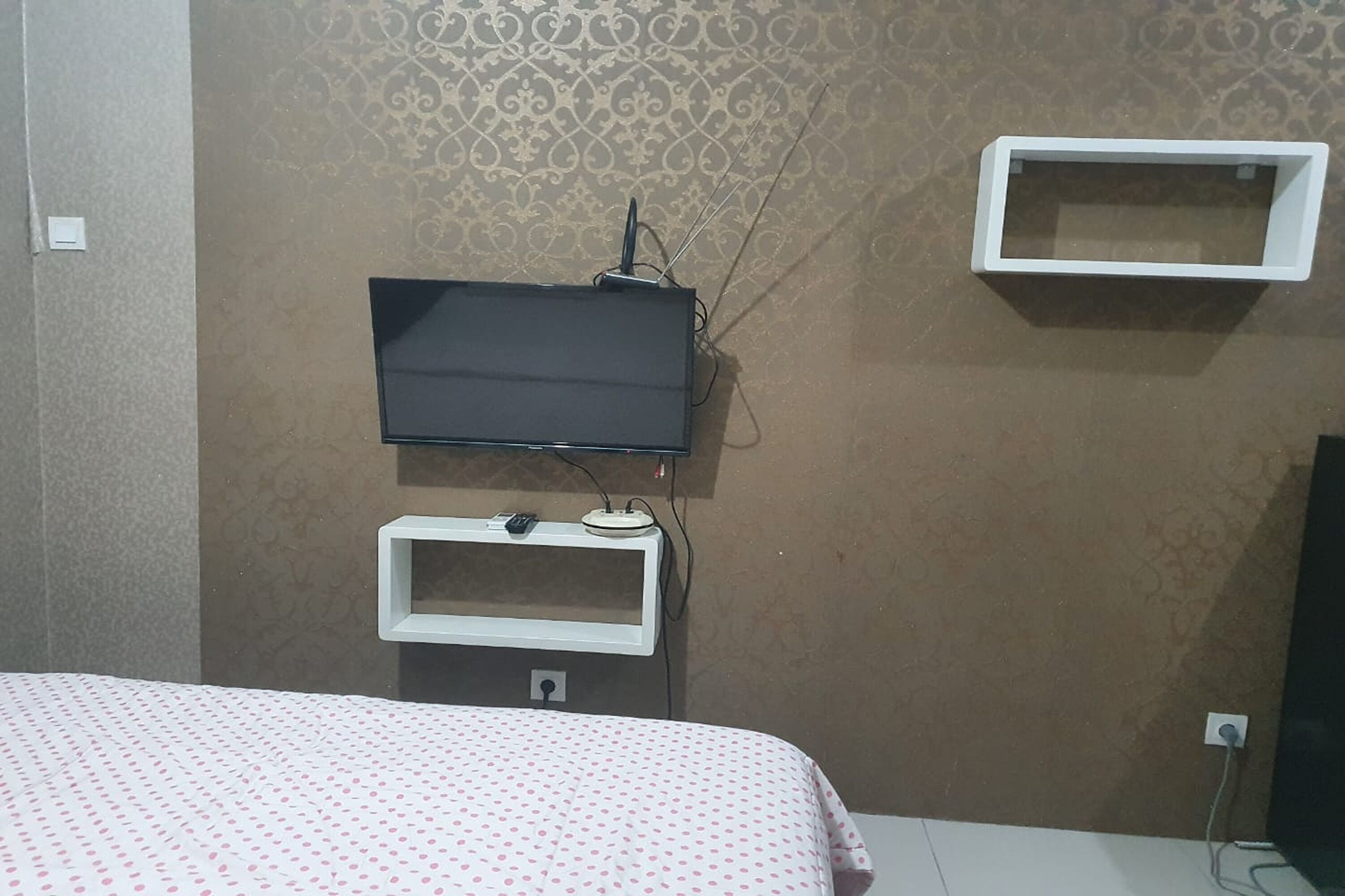 A room with a bed, a television mounted on the wall, and a small white shelf below it.