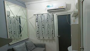 A small room with a grey sofa, a large mirror, patterned curtains, an air conditioning unit, and a door. - S&A Property 5 (Jakarta)
