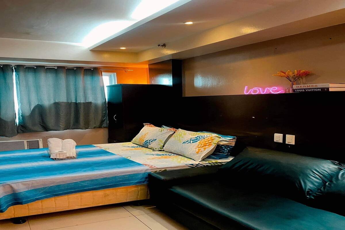 Ett modernt hotellrum med en säng, en soffa, en neonreklamskylt med ordet ”kärlek” och en vägghylla med böcker.