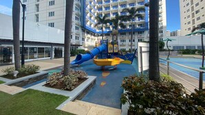 Un terrain de jeux avec un toboggan bleu et des structures de jeu jaunes, entouré de palmiers et bordé d’une piscine.