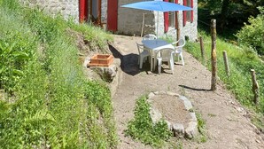 A rustic stone house with a sloped roof, red shutters, and a small porch area with a table and chairs.