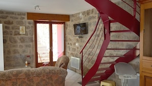 A room with a spiral staircase, a television mounted on the wall, a sofa, a chair, and a table with a floral tablecloth.