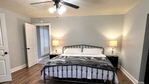 A bedroom with a bed, two bedside tables with lamps, a ceiling fan, and a door to another room.