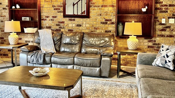 A cozy living room with a leather sofa, a wooden coffee table, and two side tables with lamps. There are built-in shelves with decorative items and a large mirror on the brick wall.
