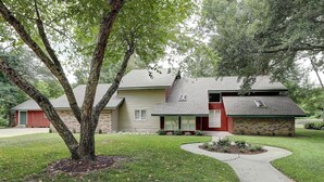 A two-story house with a brick exterior and a red door. There is a tree in the front yard and a driveway leading to the house.