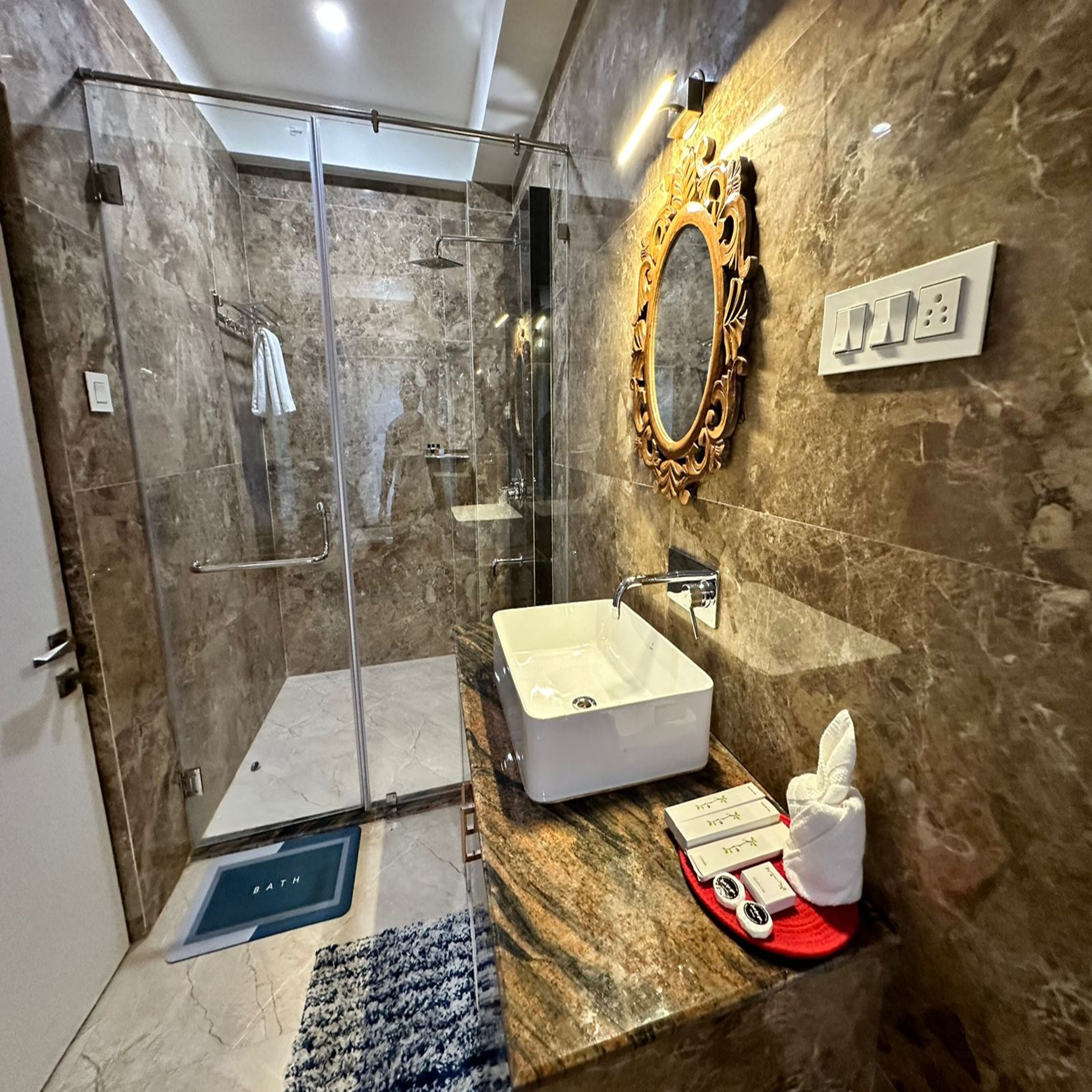 A modern bathroom with a glass-enclosed shower, a marble-tiled wall, a white sink, and a decorative mirror.