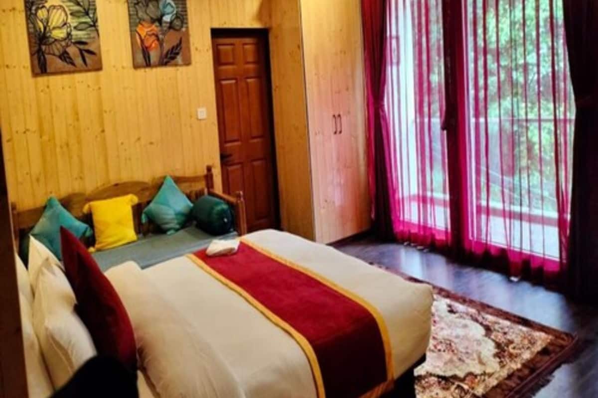 A bedroom with a bed, pillows, a wooden headboard, a window with curtains, and artwork on the wall.