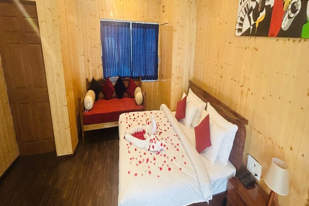 A hotel room with a wooden interior, a bed with red and white pillows, a bedside table with a lamp, and a window with blue curtains.