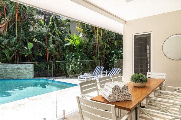 A modern outdoor pool area with lounge chairs and a table, surrounded by lush tropical plants.
