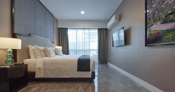 Una habitación de hotel con cama, mesita de noche, lámpara, televisión y un ventanal con cortinas.