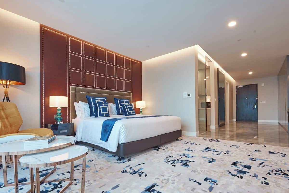 A modern hotel room with a large bed, a seating area with a chair and table, and a hallway leading to a dining area.