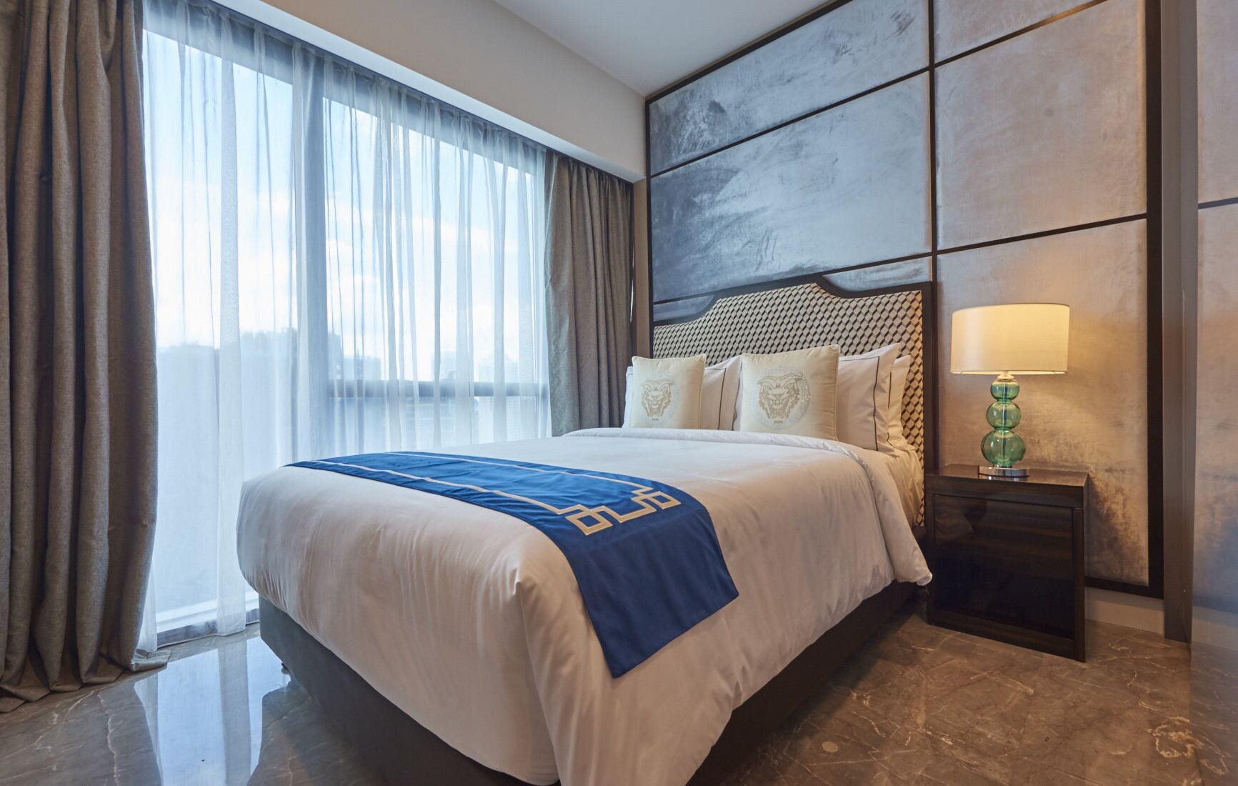 A hotel room with a large bed, a headboard with a geometric pattern, a nightstand with a lamp, and a view through sheer curtains.