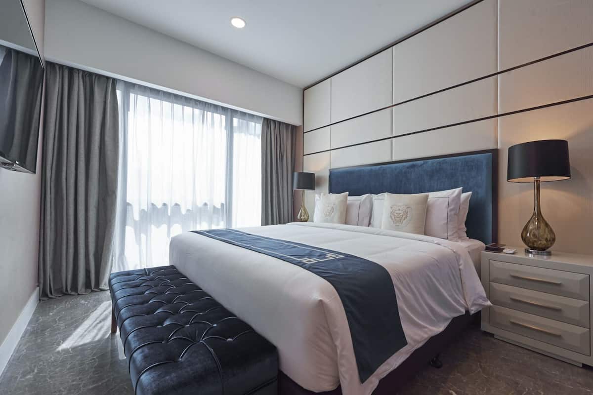 A modern hotel room with a large bed, a bedside table with a lamp, a bench, and a view of the city through large windows.