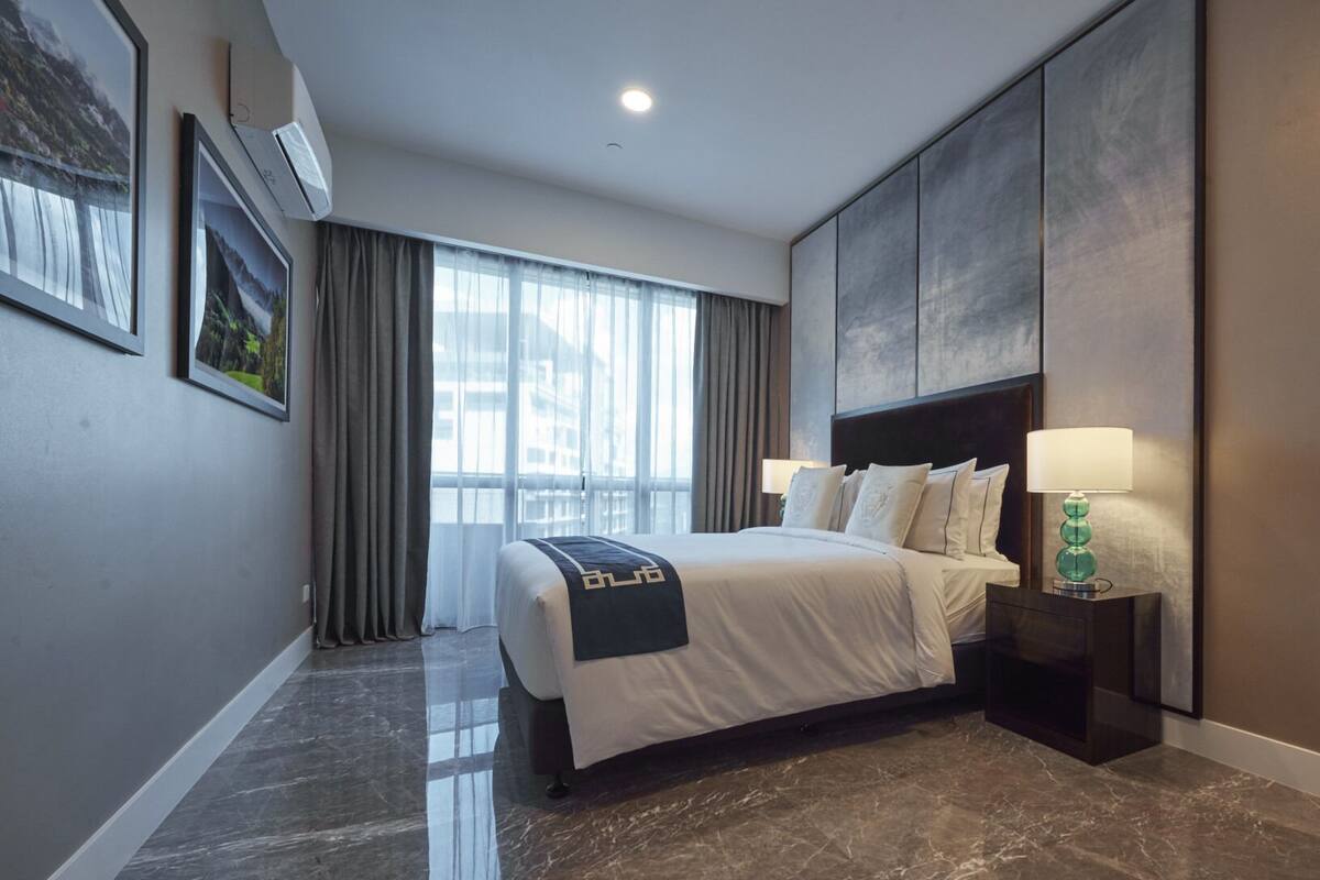 A hotel room with a large bed, a nightstand with a lamp, a window with curtains, and framed pictures on the wall.